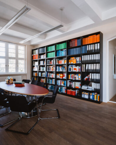 Moderner Besprechungsraum mit großem ovalem Tisch, schwarzen Stühlen, einem großen Fenster und einem hohen Regal voller Aktenordner und Fachbücher; im Hintergrund ein heller Büroflur.