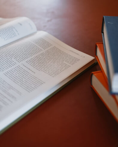 Ein aufgeschlagenes Fachbuch liegt auf einem Tisch, daneben ein Stapel geschlossener Bücher mit orangefarbenen und einem dunkelblauen Einband.