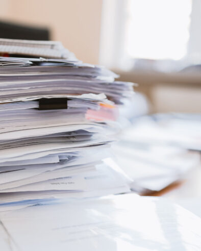 Ein hoher, chaotischer Stapel von Dokumenten und losen Blättern liegt auf einem Schreibtisch; Hintergrund unscharf mit hellem Fensterlicht.