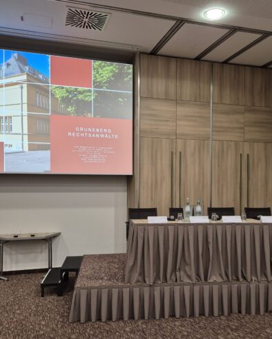 Konferenzraum mit großer Leinwand, auf der eine Willkommensfolie von ‚Gruneberg Rechtsanwälte‘ angezeigt wird. Davor ein langer Tisch für ein Podium, ausgestattet mit Wasserflaschen, Gläsern und Namensschildern.
