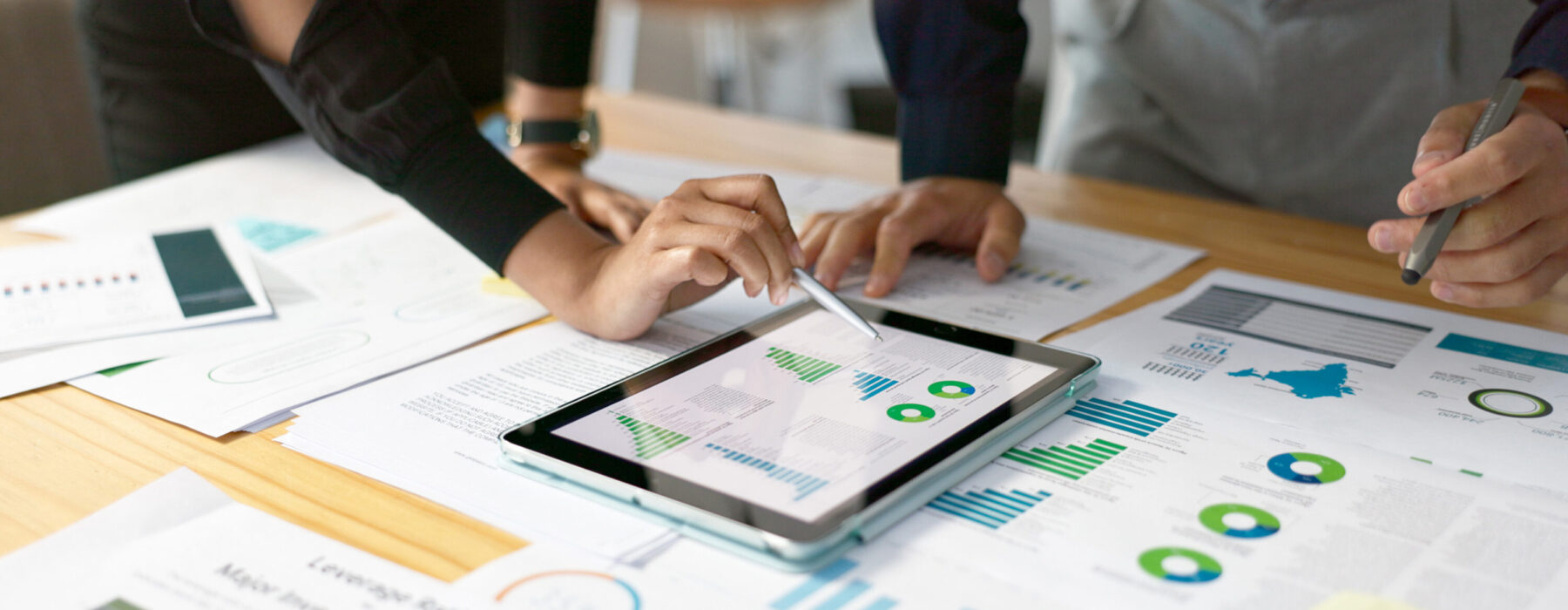 Zwei Personen analysieren Geschäftsdiagramme und Daten auf einem Tablet und ausgedruckten Berichten auf einem Schreibtisch.