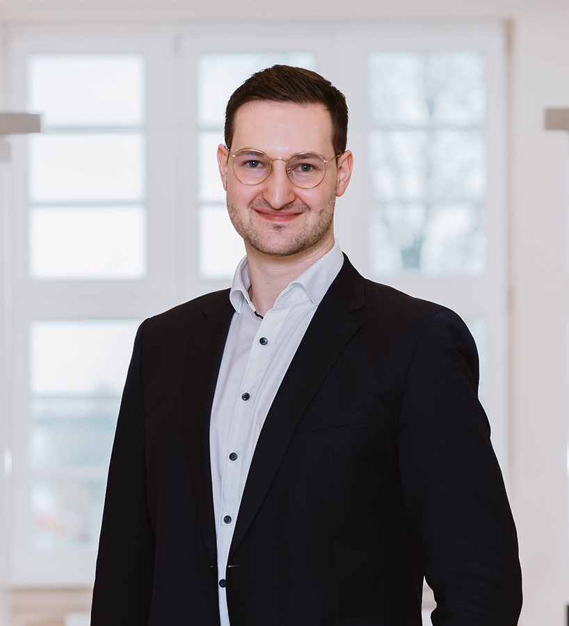 Ein junger Daniel Stausfeld mit Brille im dunklen Sakko und hellem Hemd steht vor einem großen Fenster.