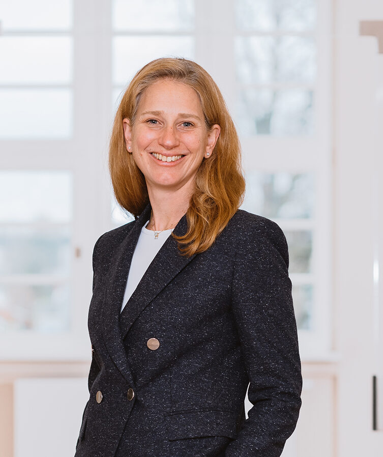 Eine lächelnde Dr. Anke Wilden-Beck mit langen Haaren in einem dunklen Blazer steht vor einem hellen Fenster.