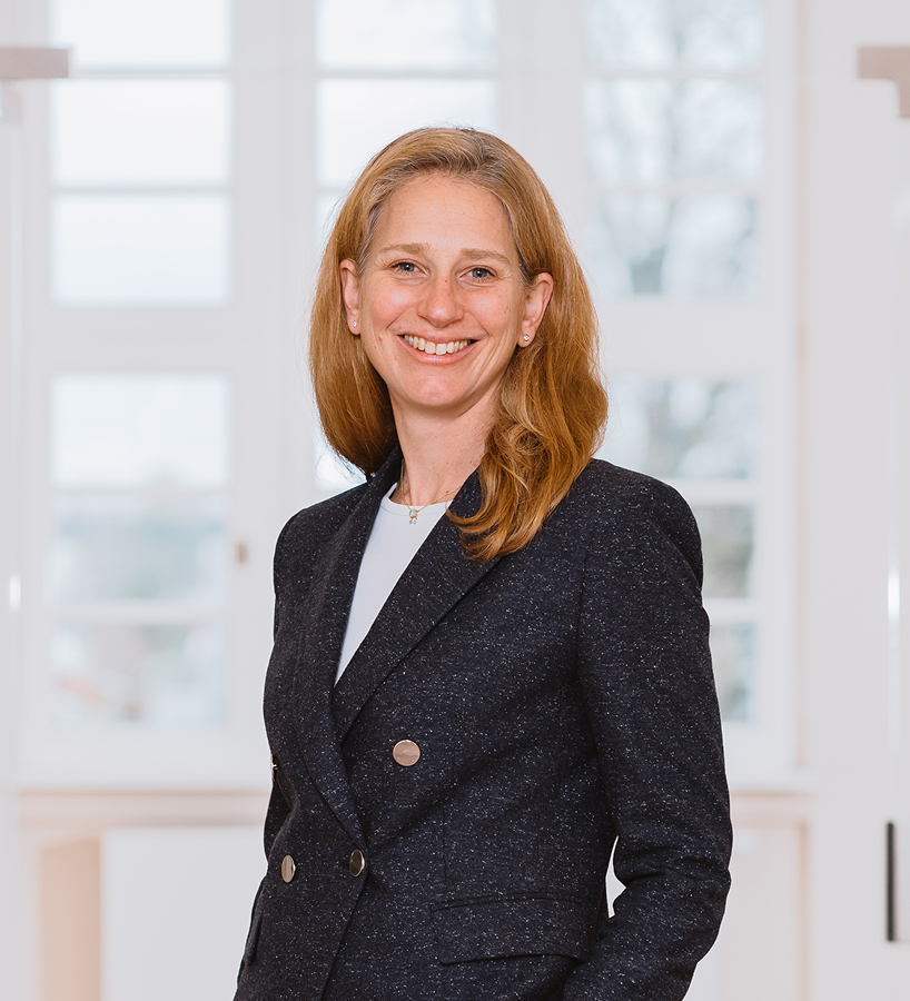 Eine lächelnde Dr. Anke Wilden-Beck mit langen Haaren in einem dunklen Blazer steht vor einem hellen Fenster.
