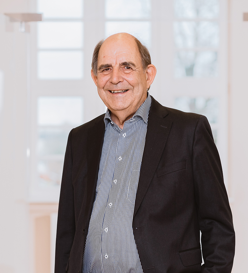 Ein älterer Dr. Ralf Bleicher im dunklen Sakko und gestreiftem Hemd lächelt vor einem hellen Fenster.