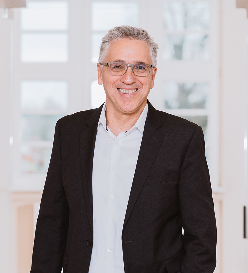 Ein lächelnder Dr. Ralf Gruneberg mit Brille in Anzug und Hemd steht vor einem hellen Fenster