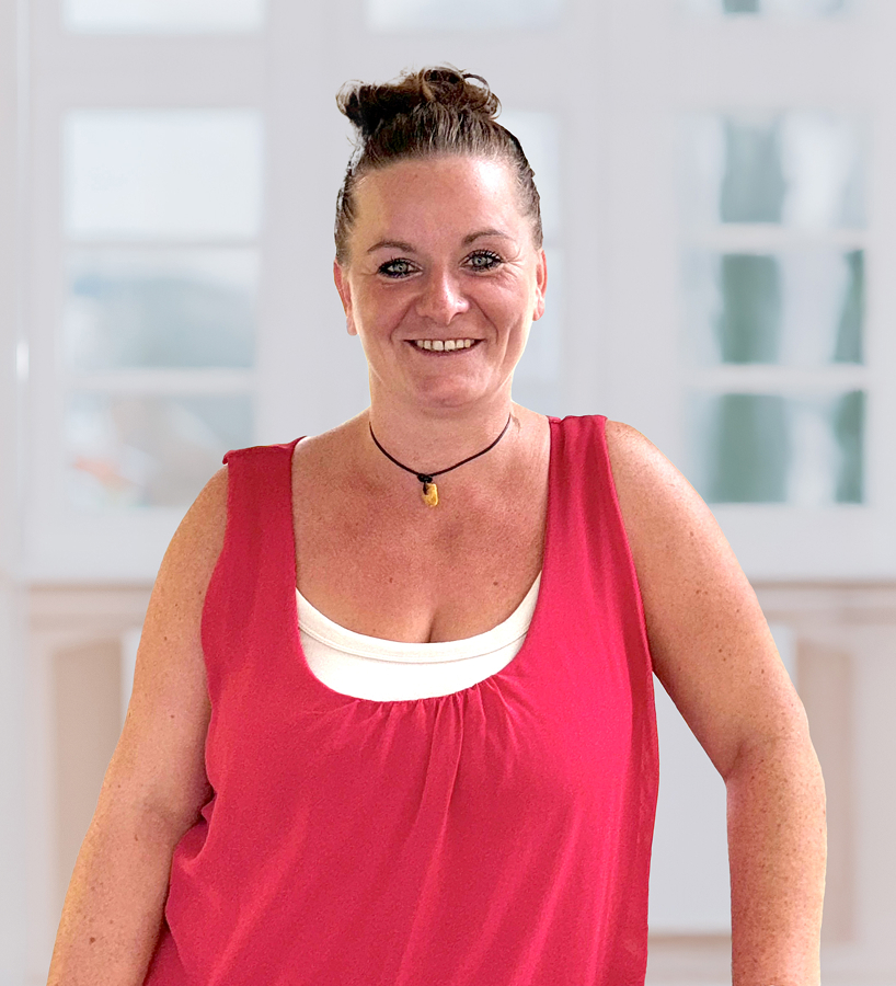 Eileen Belaschk mit hochgesteckten Haaren trägt ein rotes Oberteil und steht vor einem großen Fenster.