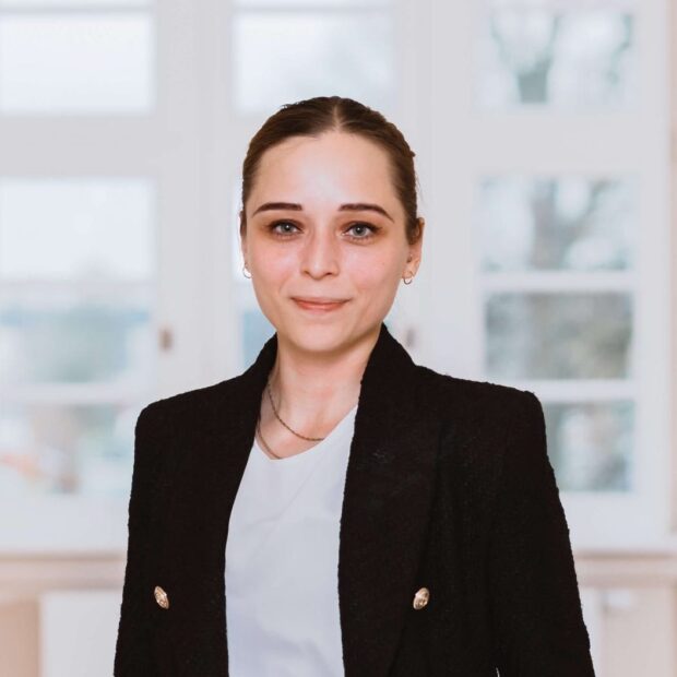 Frau in schwarzem Blazer steht lächelnd in einem hellen Büroflur.