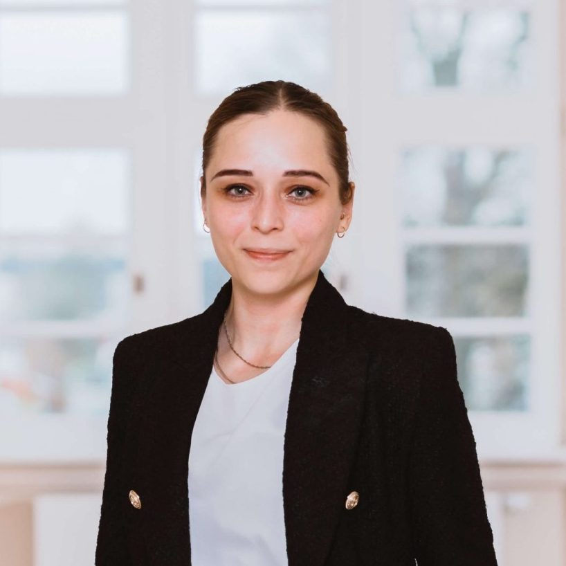 Frau in schwarzem Blazer steht lächelnd in einem hellen Büroflur.