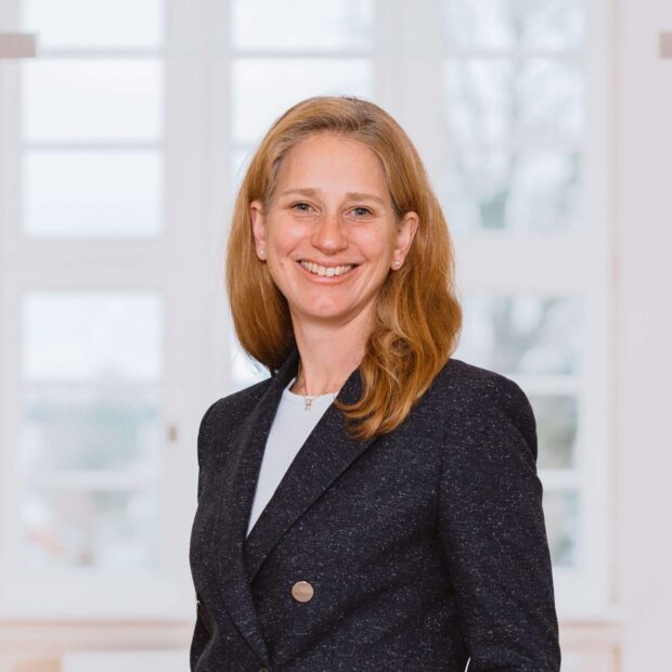 Frau in dunkelblauem Blazer steht lächelnd in einem hellen Büroflur.