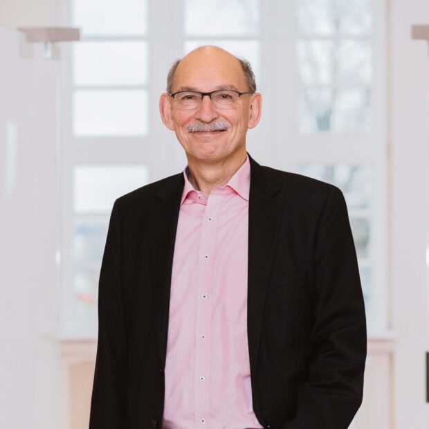 Mann in schwarzem Sakko und rosafarbenem Hemd steht lächelnd in einem hellen Büroflur.