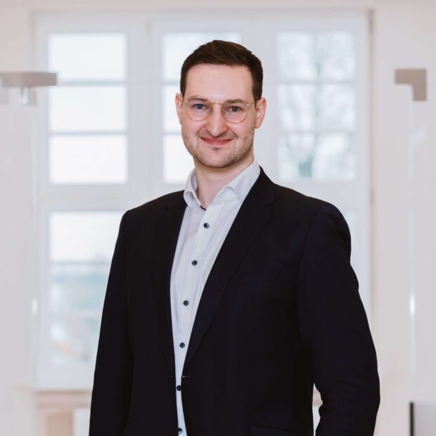 Mann im dunklen Anzug und weißem Hemd steht lächelnd in einem hellen Flur.