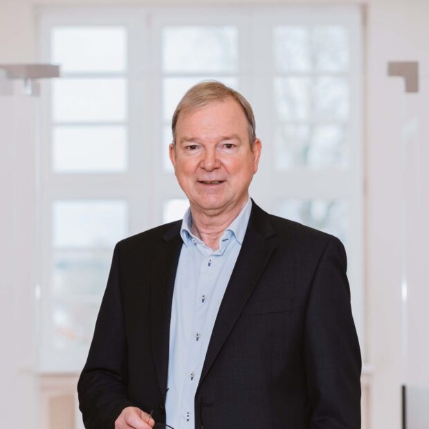 Mann in dunklem Sakko und hellem Hemd hält eine Brille und steht in einem hellen, modernen Flur.