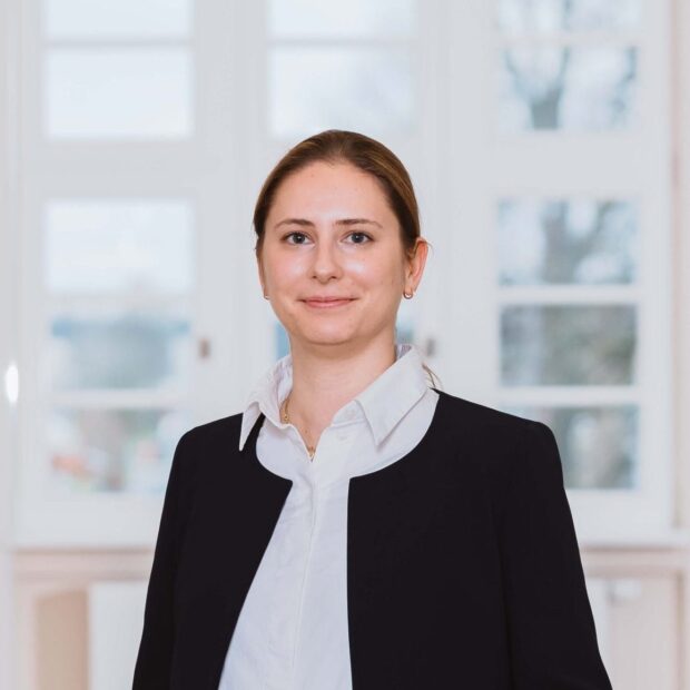 Frau in dunklem Jackett und weißer Bluse steht lächelnd in einem hellen, modernen Flur.