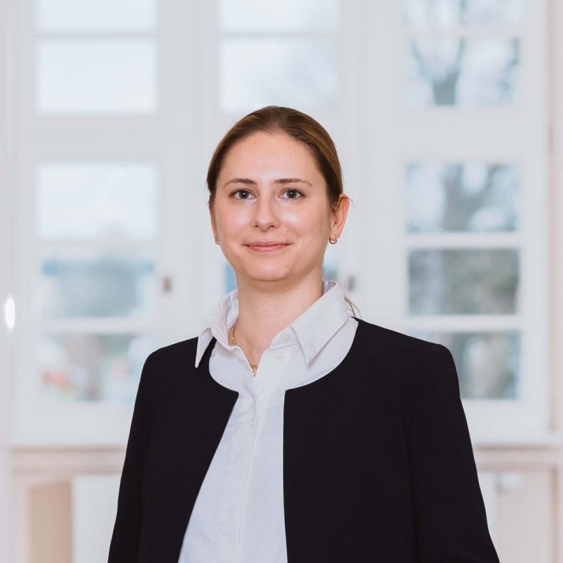 Frau in dunklem Jackett und weißer Bluse steht lächelnd in einem hellen, modernen Flur.