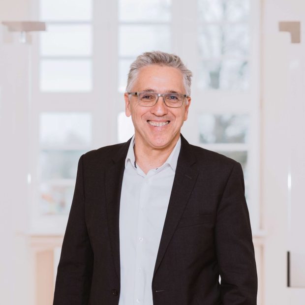 Mann mit Brille und dunklem Sakko steht lächelnd in einem hellen, modernen Flur.