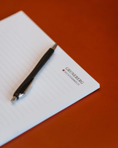 Schwarzer Kugelschreiber auf einem weißen, linierten Notizblock mit dem Logo ‚Grüneberg Rechtsanwälte‘ vor braunem Hintergrund.