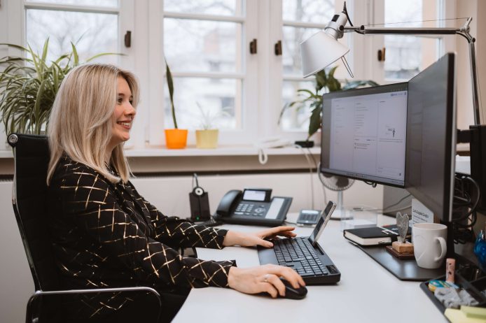 Eine Frau arbeitet lächelnd am Computer in einem Büro mit großen Fenstern.