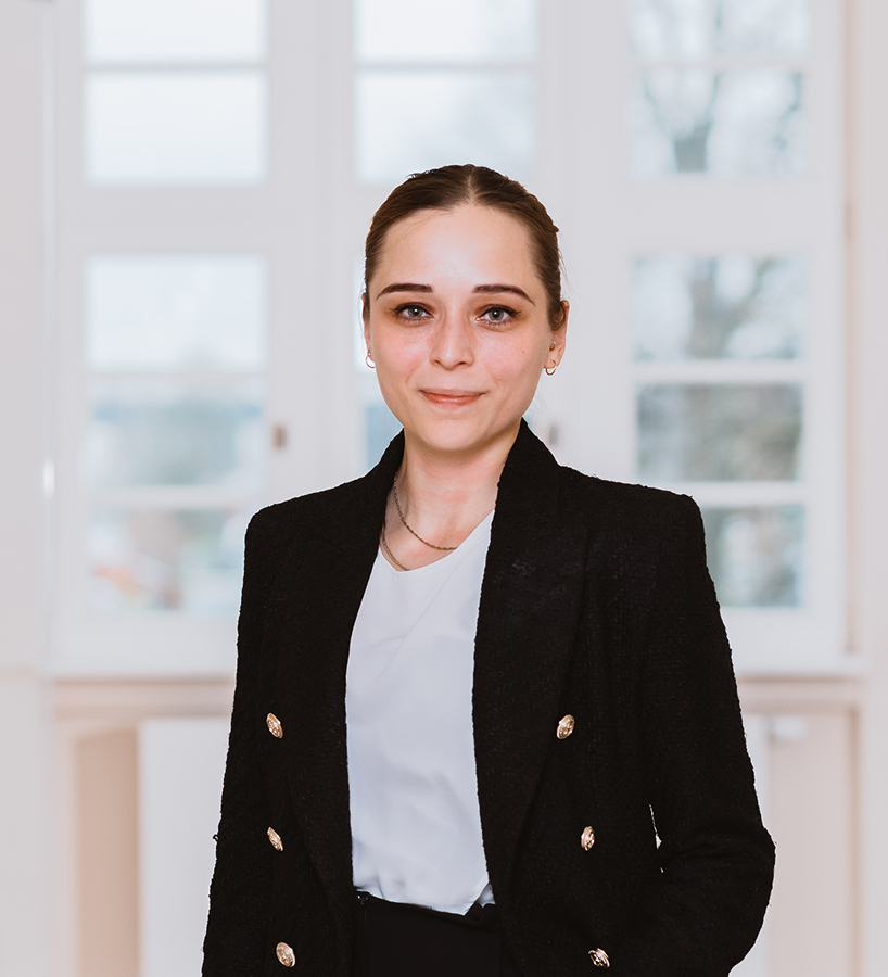 Eine junge Hannah Scholz im dunklen Blazer und hellem Oberteil steht vor einem großen Fenster und lächelt leicht.