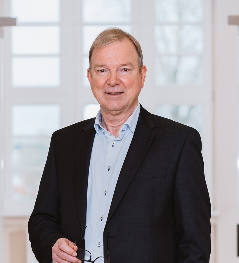 Manfred Reiche im dunklen Sakko und hellblauem Hemd hält eine Brille in der Hand und steht vor einem hellen Fenster