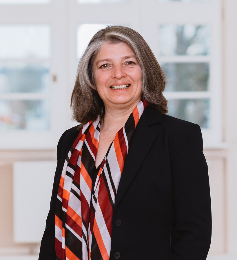 Nurşen Sengöz-Gruneberg mit schulterlangem Haar trägt einen dunklen Blazer und einen bunt gestreiften Schal, sie steht vor einem großen Fenster.