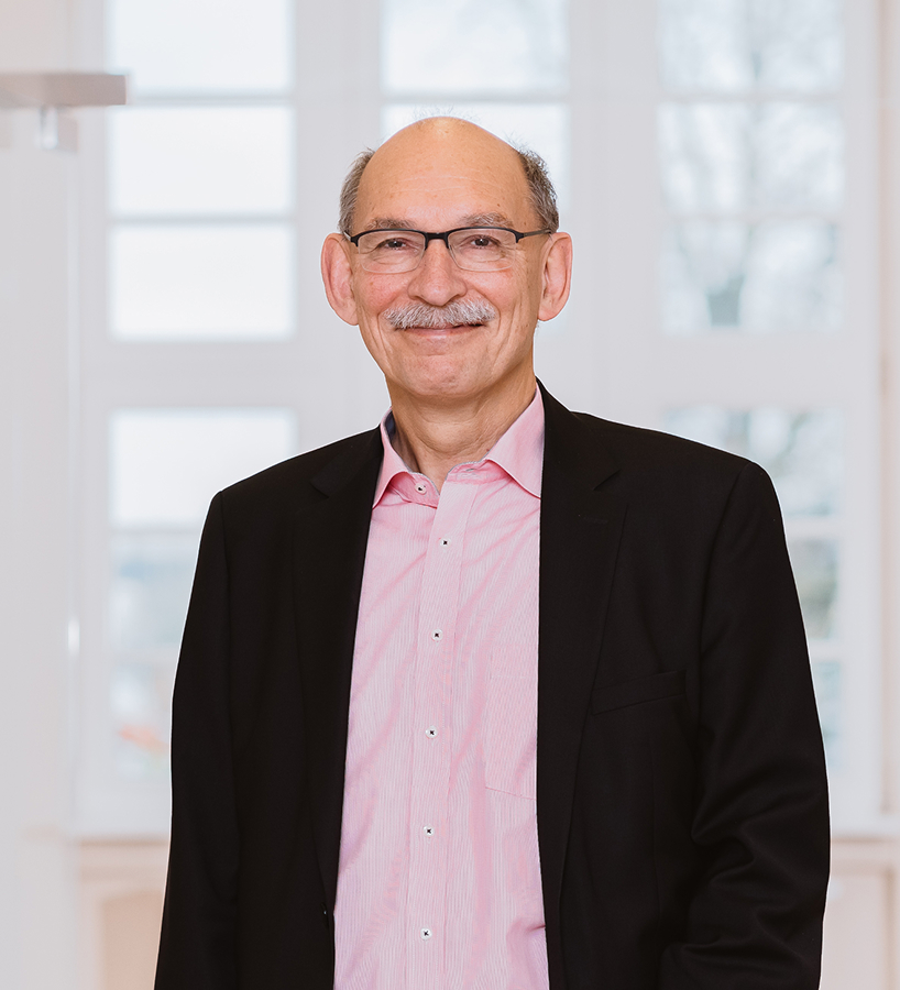 Udo Kotzea mit Brille und Schnurrbart, im dunklen Sakko und rosa Hemd, steht vor einem hellen Fenster.